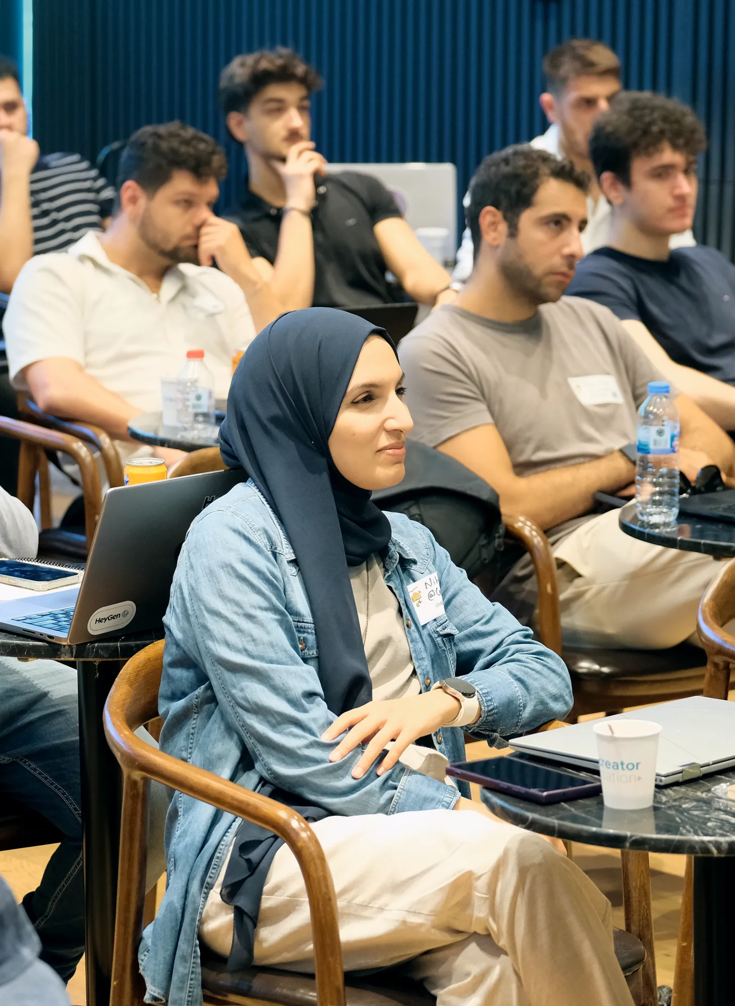 Atölye: dizüstü bilgisayarlarıyla masalarda oturan, sahneyi dinleyen katılımcılar.