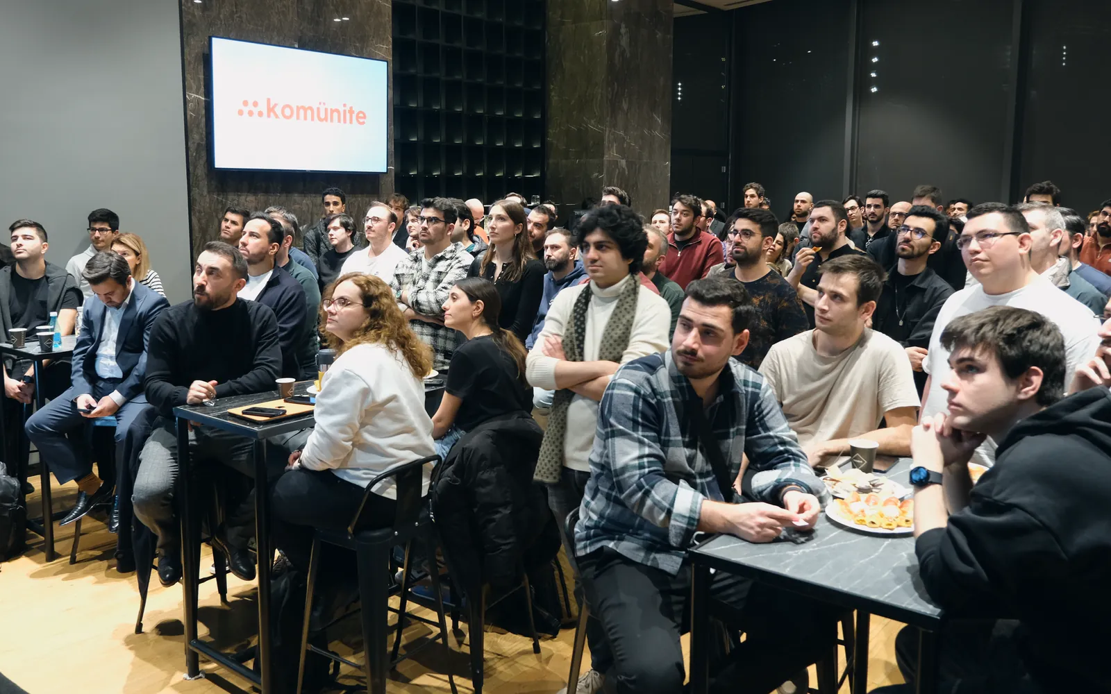 Dolu Space: ekranda Komünite logosu, masalarda ve ayakta sahneyi dinleyen kalabalık.