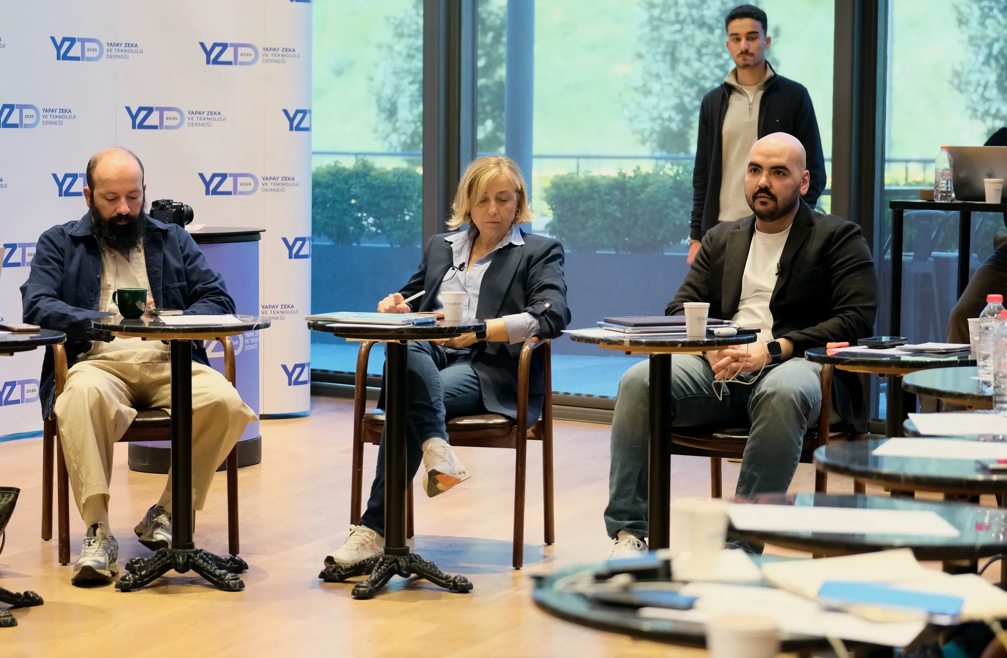 Panel: küçük mermer masaların başında oturan konuşmacılar, arkada cam cephe ve yeşillik.